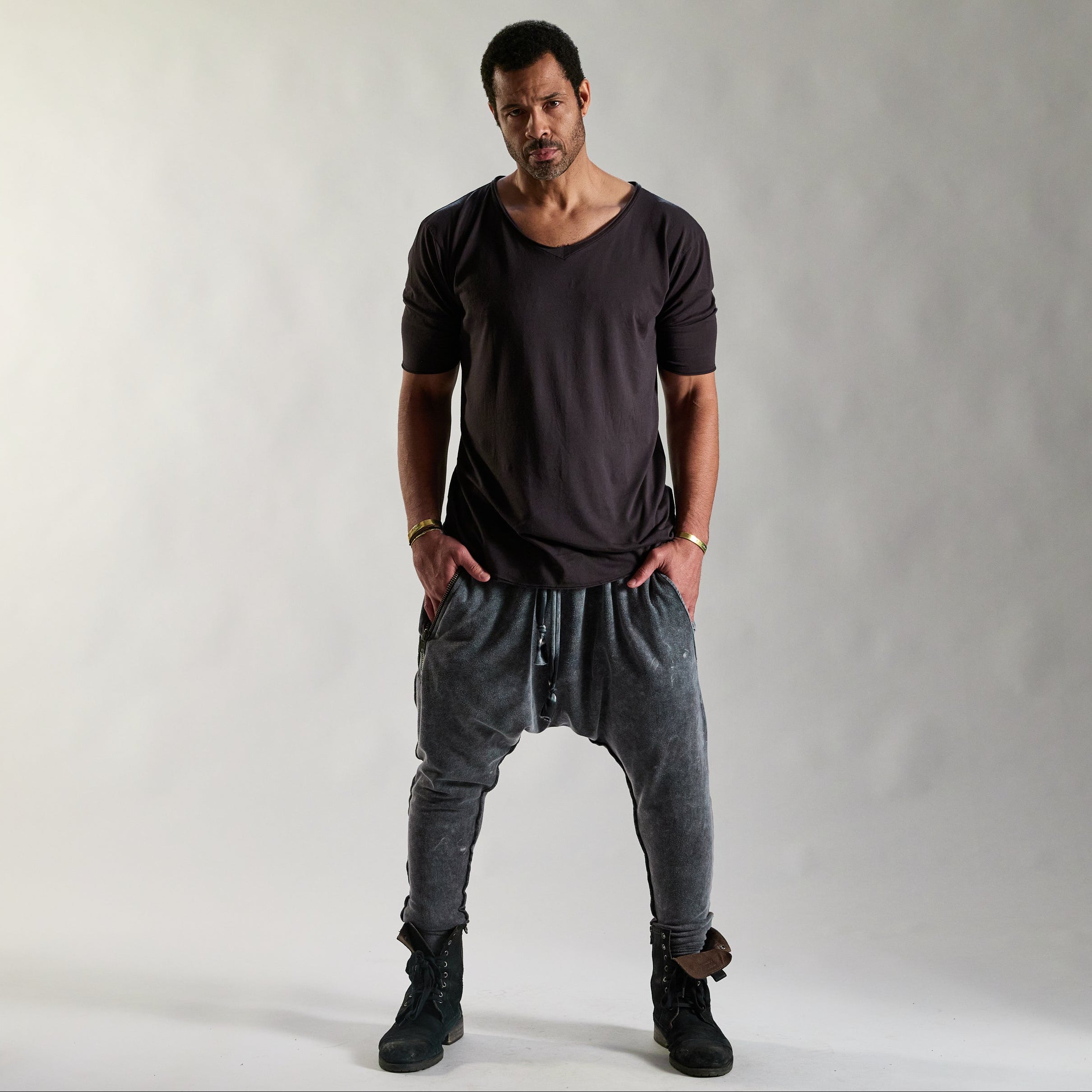 Man wearing a white long-sleeve shirt and loose grey pants, standing barefoot against a plain background.
