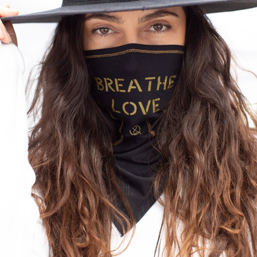 A woman wearing the One Golden Thread Versawrap as a face covering with the message "Breathe Love" in Black color