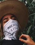 A woman wearing the Artist Collab: Gem & Bolt x One Golden Thread Versawrap as a face covering with the message "Breathe Love" in white color