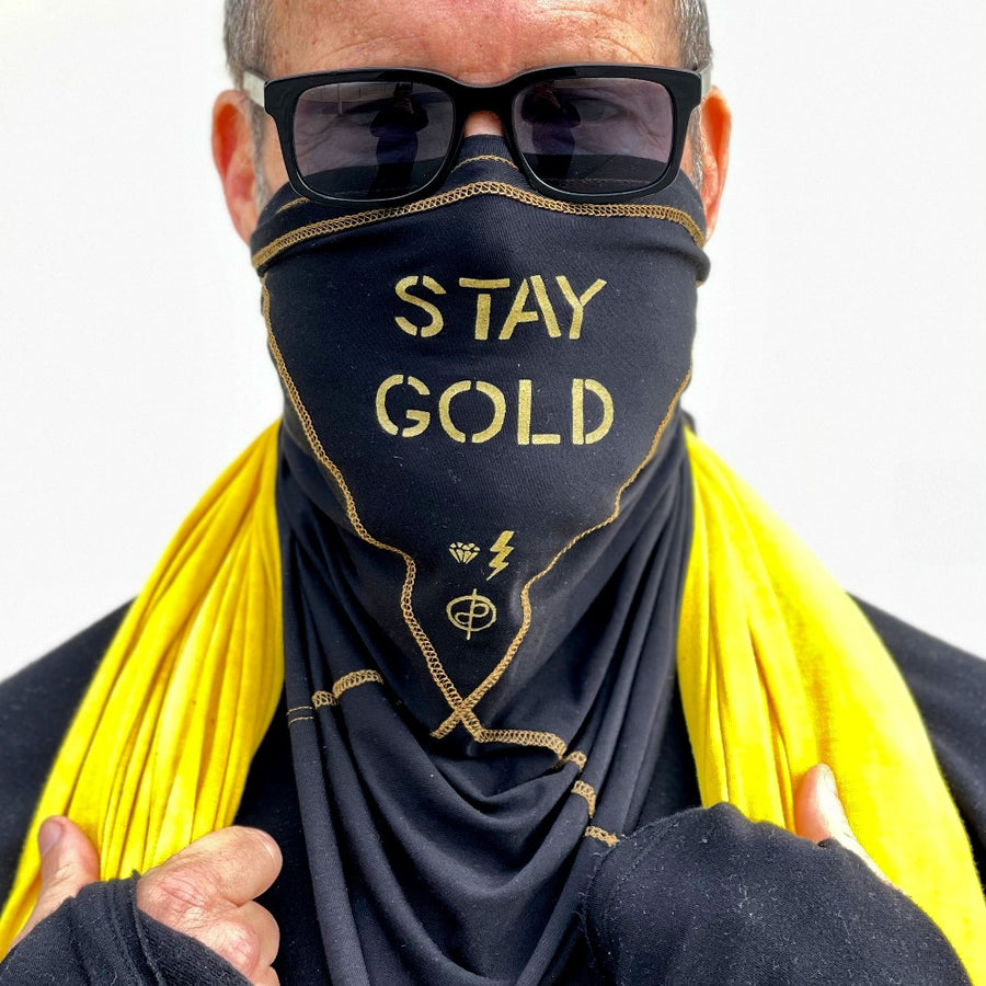 A man wearing the Artist Collab: Gem & Bolt x One Golden Thread Versawrap as a face covering with the message "Stay Gold" in black color