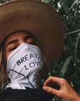 A woman wearing the One Golden Thread Versawrap as a face covering with the message "Breathe Love" in white color